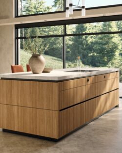 Eine moderne Team7-Kücheninsel mit Holzverkleidung und einer hellen evo-Arbeitsplatte steht in einem großzügigen Raum mit großen Fenstern und Blick auf eine grüne Außenlandschaft - perfekt, um Momente der echt.zeit zu schaffen.
