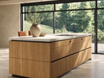 Eine moderne Team7-Kücheninsel mit Holzverkleidung und einer hellen evo-Arbeitsplatte steht in einem großzügigen Raum mit großen Fenstern und Blick auf eine grüne Außenlandschaft - perfekt, um Momente der echt.zeit zu schaffen.