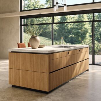 Eine moderne Team7-Kücheninsel mit Holzverkleidung und einer hellen evo-Arbeitsplatte steht in einem großzügigen Raum mit großen Fenstern und Blick auf eine grüne Außenlandschaft - perfekt, um Momente der echt.zeit zu schaffen.