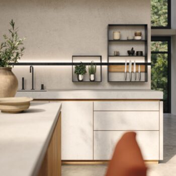 Moderne Team7 Küche mit hellen Schränken und Arbeitsflächen, Wandregalen mit Gläsern, Pflanzen und Messern sowie einem Fenster, das üppiges Grün einrahmt - perfekt, um echt.zeit Momente stilvoll zu genießen.