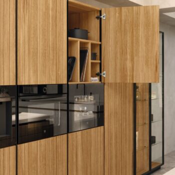 Ein moderner Team7-Küchenschrank mit vertikalen Holzpaneelen, die sich öffnen lassen, um Regale zur Aufbewahrung von Küchenutensilien und einen geschlossenen, in den Schrank eingebauten Backofen zu zeigen, präsentiert das elegante evo-Design und die zeitlose echt.zeit-Handwerkskunst.