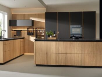 Moderne Küche mit hellen Holzschränken, schwarzen Arbeitsplatten, eingebautem Ofen und großen Fenstern, die viel Tageslicht hereinlassen.