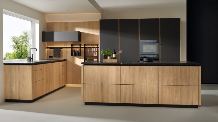 Moderne Küche mit hellen Holzschränken, schwarzen Arbeitsplatten, eingebautem Ofen und großen Fenstern, die viel Tageslicht hereinlassen.