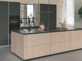 Moderne Küche mit einer Insel aus hellem Holz, schwarzer Arbeitsplatte, dunklen Schränken, Einbaugeräten, einem Messerblock und einer Topfpflanze in der Ecke.