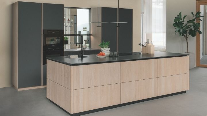 Moderne Küche mit einer Insel aus hellem Holz, schwarzer Arbeitsplatte, dunklen Schränken, Einbaugeräten, einem Messerblock und einer Topfpflanze in der Ecke.