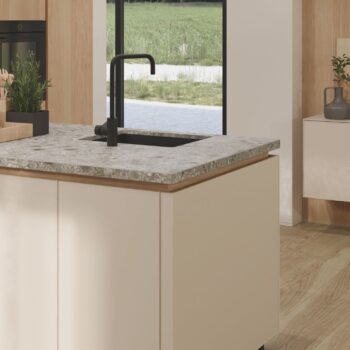 Eine moderne Küche mit einer Arbeitsplatte aus reinem Marmor, einem schwarzen Wasserhahn, beigen Schränken und einem großen Fenster, das den Blick auf einen grasbewachsenen Außenbereich freigibt - eine perfekte Verkörperung der eleganten Designphilosophie von Team7.