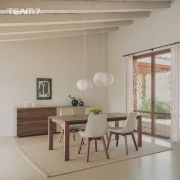 Ein modernes Esszimmer mit einem Team 7 Auszugstisch aus Holz, vier gepolsterten Stühlen, zwei Pendelleuchten, einem Sideboard, Kunstwerken und großen Glastüren.