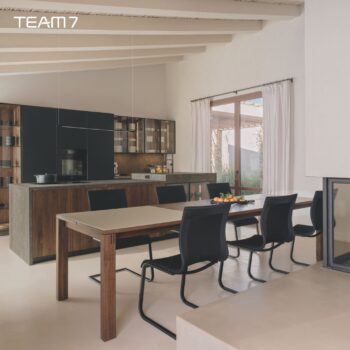 Moderne Küche und Essbereich mit Team 7 Holzschränken, einer großen zentralen Insel, einem langen Auszugstisch mit sechs schwarzen Stühlen, großem Fenster mit Vorhängen und minimalistischem Dekor.