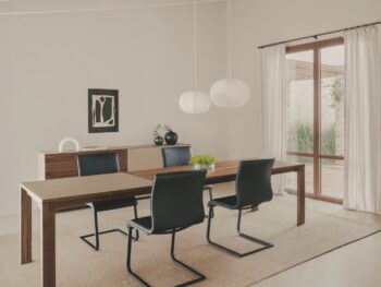 Modernes Esszimmer mit einem Team 7 Auszugstisch aus Holz, vier schwarzen Stühlen, einem Sideboard, zwei Pendelleuchten und großen Fenstern mit transparenten Vorhängen. Minimalistische Dekoration mit Kunstwerken und Vasen für einen eleganten Magnum-Look.