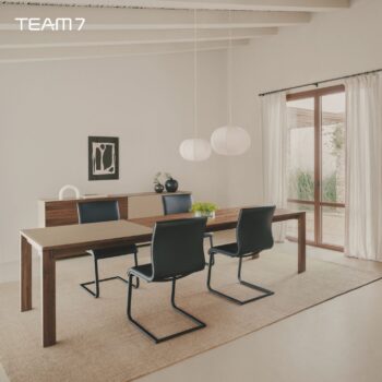 Modernes Esszimmer mit einem Team 7 Auszugstisch aus Holz, vier schwarzen Stühlen, einem Sideboard, zwei Pendelleuchten und großen Fenstern mit transparenten Vorhängen. Minimalistische Dekoration mit Kunstwerken und Vasen für einen eleganten Magnum-Look.