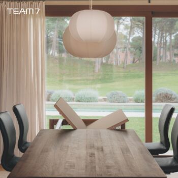 Ein modernes Esszimmer mit einem TEAM 7 Auszugstisch aus Holz, schwarzen Stühlen, einer weißen Hängeleuchte und großen Fenstern mit Blick auf einen grünen Rasen und Bäume. Das Wort TEAM 7 erscheint in der oberen linken Ecke.