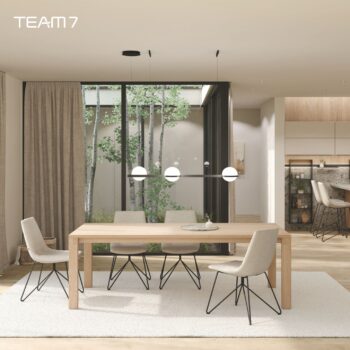 Ein modernes Esszimmer mit einem Team 7 magnum Auszugstisch aus hellem Holz, vier beigen Stühlen, einem weißen Teppich und Pendelleuchten. Große Fenster geben den Blick auf grüne Bäume frei, im Hintergrund ist der Küchenbereich zu sehen.