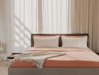Ein modernes Team7 Bett Riletto mit pfirsichfarbener Bettwäsche, zwei weißen Kissen und einer grauen Überwurfdecke steht vor hohen, transparenten Vorhängen. Das TEAM7-Logo erscheint in der oberen linken Ecke.