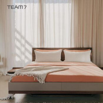 Ein modernes Team7 Bett Riletto mit pfirsichfarbener Bettwäsche, zwei weißen Kissen und einer grauen Überwurfdecke steht vor hohen, transparenten Vorhängen. Das TEAM7-Logo erscheint in der oberen linken Ecke.