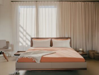 Modernes Schlafzimmer mit einem Team7 Bett Riletto-Bettgestell aus Holz, orangefarbener Bettwäsche, zwei weißen Kissen, einem Stuhl links und Körben rechts, vor durchsichtigen Vorhängen und beigen Wänden.