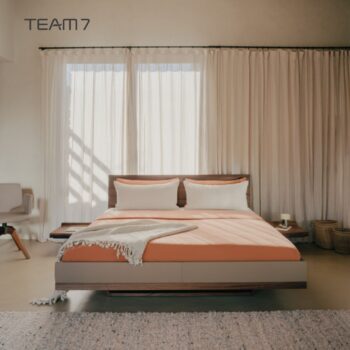 Modernes Schlafzimmer mit einem Team7 Bett Riletto-Bettgestell aus Holz, orangefarbener Bettwäsche, zwei weißen Kissen, einem Stuhl links und Körben rechts, vor durchsichtigen Vorhängen und beigen Wänden.