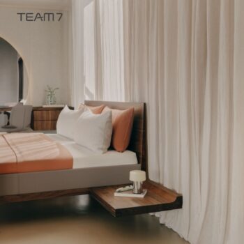 Modernes Schlafzimmer mit dem Team7 Bett Riletto, weißem und orangefarbenem Bettzeug, einem schwebenden Nachttisch mit Lampe und Buch, hellen Vorhängen und dem TEAM 7 Logo an der Wand.
