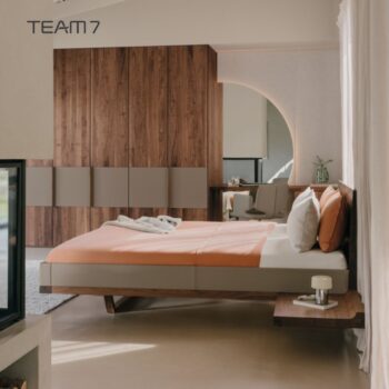 Modernes Schlafzimmer mit dem Bett Riletto-Holzbettgestell von Team7, orangefarbener Bettwäsche, einem Einbauschrank aus Holzpaneelen und einem minimalistischen Waschtisch mit einem runden Spiegel. Der Raum ist von natürlichem Licht durchflutet.