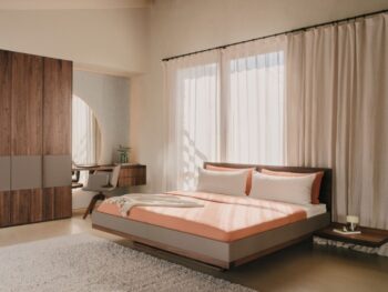 Ein modernes Schlafzimmer mit einem Bett Riletto von Team7, einem Holzbett, orangefarbener Bettwäsche, zwei Kissen, einem grauen Teppich, hölzernen Nachttischen, einem großen Kleiderschrank und transparenten Vorhängen, die natürliches Licht hereinlassen.
