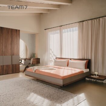 Ein modernes Schlafzimmer mit einem Bett Riletto von Team7, einem Holzbett, orangefarbener Bettwäsche, zwei Kissen, einem grauen Teppich, hölzernen Nachttischen, einem großen Kleiderschrank und transparenten Vorhängen, die natürliches Licht hereinlassen.