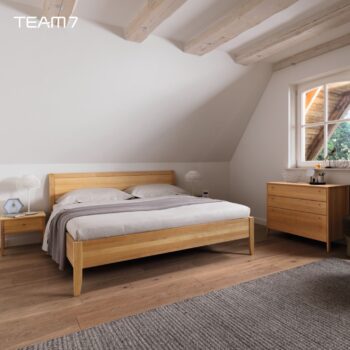 Ein modernes Schlafzimmer mit einem Holzbett, passenden Nachttischen und einer Kommode, neutraler Bettwäsche, einem grauen Teppich und einem Fenster, das natürliches Licht hereinlässt, schafft eine warme, von Sesam inspirierte Atmosphäre.