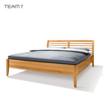 Ein hölzernes Sesam-Bettgestell mit Lattenrost, hergestellt von TEAM 7, abgebildet mit einer grauen Bettdecke und einem Kissen vor weißem Hintergrund.