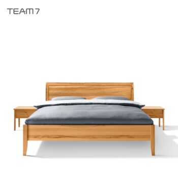 Ein hölzernes Sesam-Bett mit Kopfteil, grauer Bettwäsche und zwei passenden hölzernen Nachttischen, alles vor einem weißen Hintergrund. Der Markenname TEAM7 erscheint in der oberen linken Ecke.