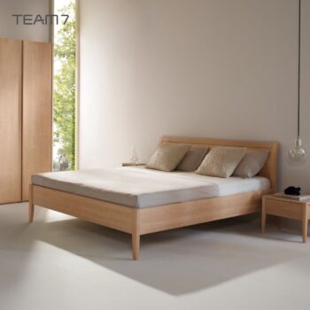 Minimalistisches Schlafzimmer mit einem Bettgestell aus Sesamholz, hellgrauer Bettwäsche, zwei beigen Kissen, einem kleinen hölzernen Nachttisch und einer Hängelampe neben einem großen Fenster.