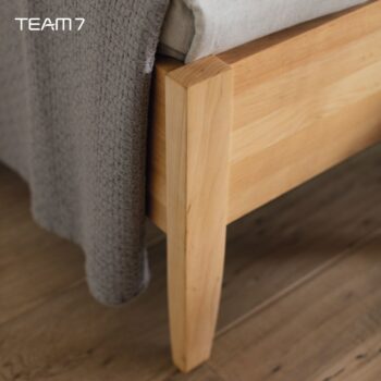 Nahaufnahme eines Bettgestells aus Sesamholz, über das eine graue, strukturierte Decke drapiert ist, die auf einem Holzboden liegt. Das TEAM 7-Logo erscheint in der oberen linken Ecke.