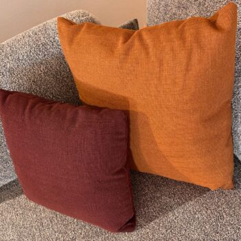 Zwei quadratische Zierkissen – eines in gebranntem Orange und eines in Dunkelrot – liegen auf einem grauen Vitra-Sofa mit strukturierter Oberfläche. Die auffälligen Farben sorgen in Ihrem Wohnbereich für eine einladende Mischung aus Wärme und Stil.