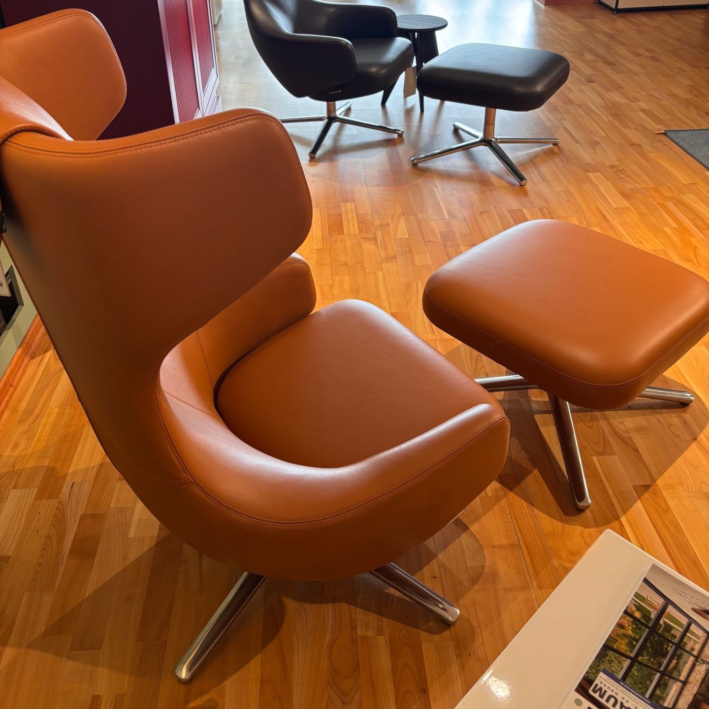 Brauner Ledersessel mit passendem Hocker auf einem Holzboden, im Hintergrund ein weiterer schwarzer Vitra-Loungesessel mit Hocker. In der rechten unteren Ecke ist ein Teil einer Zeitschrift zu sehen, was die einladende Atmosphäre unterstreicht, die an eine klassische „Grand Repos“-Kulisse erinnert. Brauner Ledersessel mit passendem Hocker auf einem Holzboden, im Hintergrund ein weiterer schwarzer Vitra-Loungesessel mit Hocker. In der rechten unteren Ecke ist ein Teil einer Zeitschrift zu sehen, was die einladende Atmosphäre unterstreicht, die an eine klassische „Grand Repos“-Kulisse erinnert.