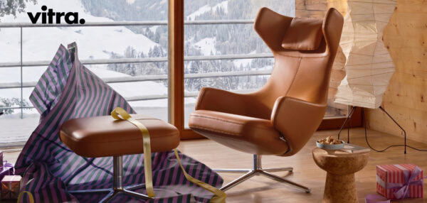 Ein brauner Ledersessel mit passendem Fußhocker, der teilweise mit gestreiftem Papier umwickelt ist, steht vor einem Fenster mit Blick auf verschneite Berge – eine perfekte Szene, die vom Geist der „Vitra Home Stories Winter 2026/2027“ inspiriert ist. Verpackte Geschenke und eine Lampe in der Nähe tragen zur einladenden Atmosphäre bei und unterstreichen gutes Design bis ins kleinste Detail.