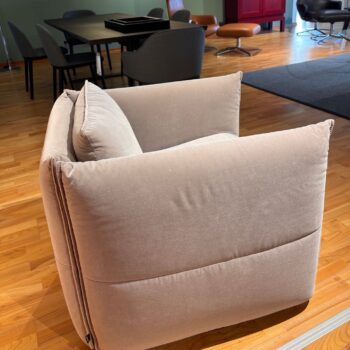 Ein beiger, gepolsterter Vitra-Sessel auf einem Holzboden in einem modernen Raum mit Tischen, Stühlen und einem roten Schrank im Hintergrund, der das elegante Ambiente des Mariposa Clubs heraufbeschwört.