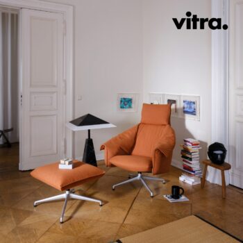 Ein orangefarbener Vitra-Lounge-Sessel mit passendem Hocker in einem Raum mit Holzboden, einem Beistelltisch, Büchern, einer Lampe und Kunstwerken an den Wänden. Das Wort „vitra.“ befindet sich in der oberen rechten Ecke.