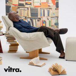 Ein Mann lehnt in einem beigen Vitra-Sessel mit Ottomane, umgeben von verstreuten Büchern, vor einer Wand aus Bücherregalen. In der unteren linken Ecke ist das Wort „vitra.“ zu sehen.
