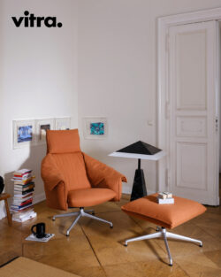 Ein modernes Wohnzimmer mit einem orangefarbenen Sessel und einer dazu passenden Ottomane, einem schwarzen Tisch mit Büchern, gerahmten Kunstwerken an der Wand und Büchern, die neben einer weißen Tür gestapelt sind, verkörpert die Essenz von gutem Design. Das Vitra-Logo ist in der Ecke zu sehen und hebt diese einladende Szene als Teil der „Vitra Home Stories“ für den Winter 2026/2027 hervor.