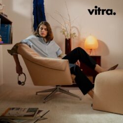 Eine Person sitzt entspannt in einem beigen Vitra-Drehsessel in einem modernen Wohnzimmer, im Hintergrund sind Bücher, eine Lampe, Vasen und das Vitra-Logo zu sehen – dies schafft ein stilvolles Ambiente, das die zeitlose Eleganz des Bascule-Designs widerspiegelt.