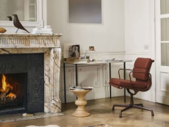 Ein Heimbüro mit Marmorkamin, beleuchtetem Feuer, modernem Schreibtisch und Vitra Soft Pad Chair EA 205, rundem Holzhocker, verstreuten Zeitschriften auf dem Boden und minimalem Dekor in einem hellen Raum.
