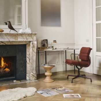 Ein Heimbüro mit Marmorkamin, beleuchtetem Feuer, modernem Schreibtisch und Vitra Soft Pad Chair EA 205, rundem Holzhocker, verstreuten Zeitschriften auf dem Boden und minimalem Dekor in einem hellen Raum.