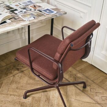 Ein kastanienbrauner Vitra Soft Pad Chair mit Armlehnen und drehbarem Untergestell steht neben einem Schreibtisch mit Marmorplatte, auf dem aufgeschlagene Zeitschriften und Papiere auf einem Parkettboden liegen.
