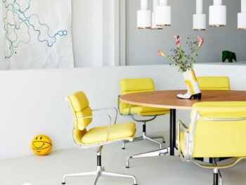 Ein runder Holztisch mit Vitra Soft Pad Chairs, eine Vase mit Blumen, weiße Pendelleuchten, abstrakte Wandkunst und ein gelber Basketball mit einem Smiley-Gesicht.