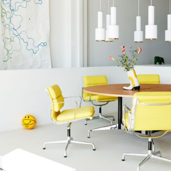 Ein runder Holztisch mit Vitra Soft Pad Chairs, eine Vase mit Blumen, weiße Pendelleuchten, abstrakte Wandkunst und ein gelber Basketball mit einem Smiley-Gesicht.