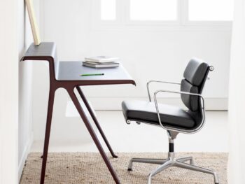 Ein moderner, minimalistischer Schreibtisch mit einem Vitra Soft Pad Chair EA 207 auf einem gewebten Teppich; auf dem Schreibtisch liegen Bücher, ein Notizbuch und ein Bleistift, im Hintergrund ein Fenster.