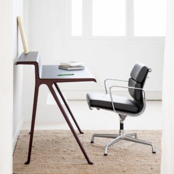 Ein moderner, minimalistischer Schreibtisch mit einem Vitra Soft Pad Chair EA 207 auf einem gewebten Teppich; auf dem Schreibtisch liegen Bücher, ein Notizbuch und ein Bleistift, im Hintergrund ein Fenster.