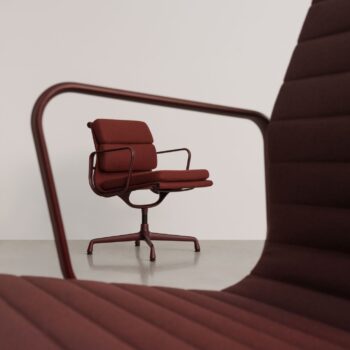 Im Hintergrund ist ein kastanienbrauner, gepolsterter Vitra Soft Pad Chair mit Metallgestell und Drehfuß zu sehen, eingerahmt von den Arm- und Rückenlehnen eines passenden Stuhls im Vordergrund.