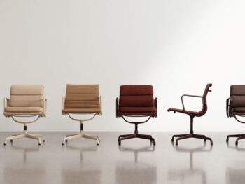Fünf moderne Drehstühle, darunter der ikonische Vitra Soft Pad Chair EA 219, in neutralen und rötlich-braunen Tönen sind in einer Reihe vor einer schlichten weißen Wand auf einem polierten Boden angeordnet.