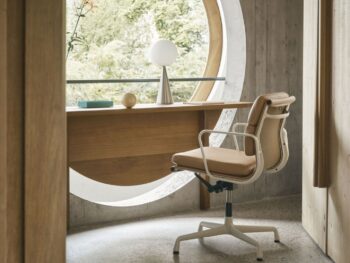 In einem modernen Heimbüro steht ein Vitra Soft Pad Chair an einem Holzschreibtisch vor einem großen runden Fenster mit Blick ins Grüne. Zur minimalistischen Einrichtung gehören eine Lampe und eine Schale auf dem Schreibtisch.