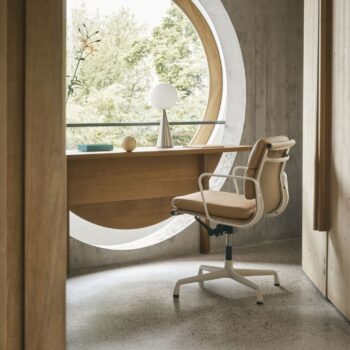In einem modernen Heimbüro steht ein Vitra Soft Pad Chair an einem Holzschreibtisch vor einem großen runden Fenster mit Blick ins Grüne. Zur minimalistischen Einrichtung gehören eine Lampe und eine Schale auf dem Schreibtisch.