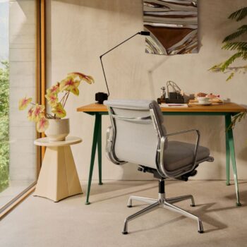 Ein modernes Heimbüro mit einem Vitra Soft Pad Chair, einem Holzschreibtisch mit grünen Beinen, Büromaterial, einer Topfpflanze auf einem beigen Ständer und einer Wandlampe am Fenster.