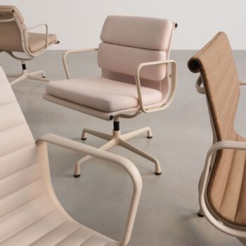 Vier moderne Vitra Soft Pad Chairs, darunter die Modelle EA 205 und EA 207, mit gepolsterten Sitzen und Metallarmlehnen sind auf einem glatten, hellgrauen Boden vor einer schlichten weißen Wand angeordnet.